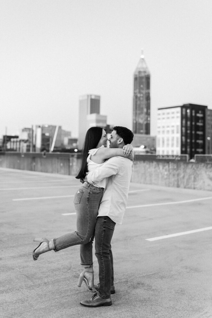 Man picks up woman during cityscape engagement in Atlanta, GA. Atlanta skyline view is in the background.