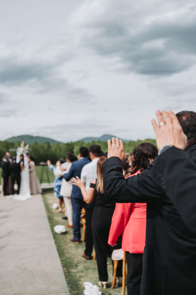 Guests raise hands and pray over bride and groom during ceremony. Wedding at Meadows at Mossy Creek.
