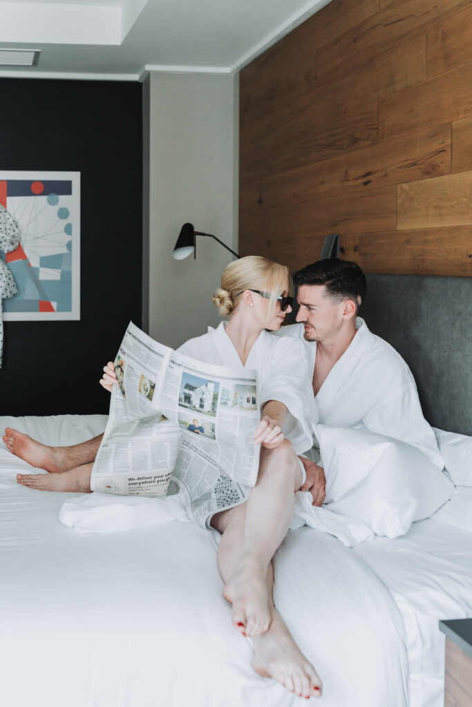 Bride and groom sit on hotel bed in their robes with a newspaper before their city elopement at epicurean hotel atlanta