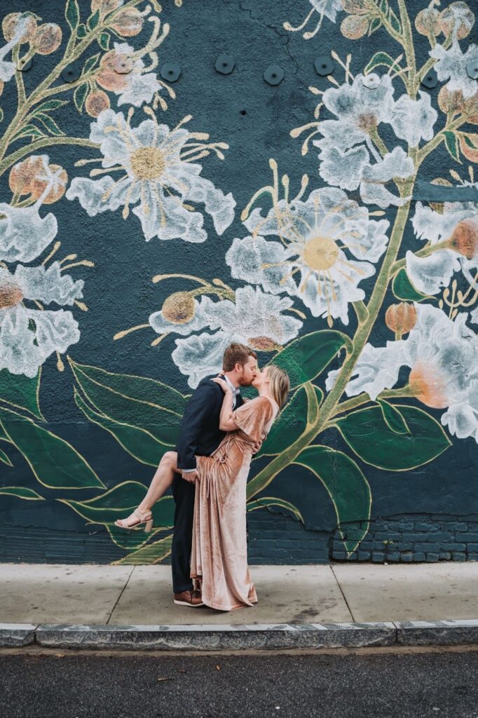 An engaged couple kissing on a sidewalk if front of a wall