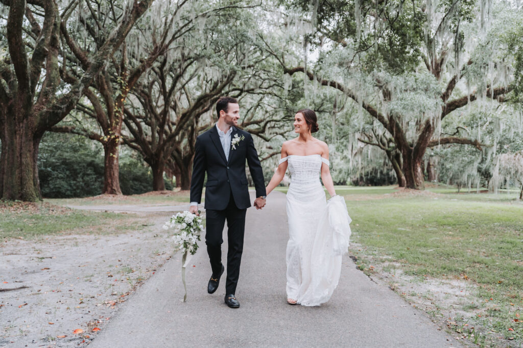 Bride and groom hold hands and walk under oak trees at Legare waring House wedding venue.