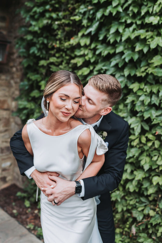 groom kisses bride's cheek while he holds her from behind