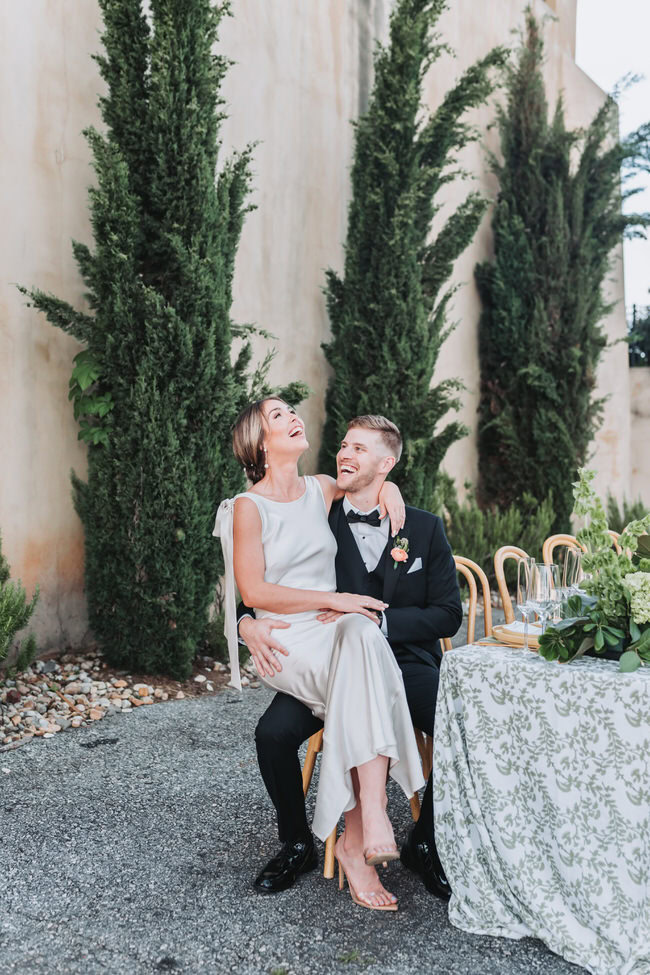 bride laughs while sitting in the groom's lap at the wedding reception table.