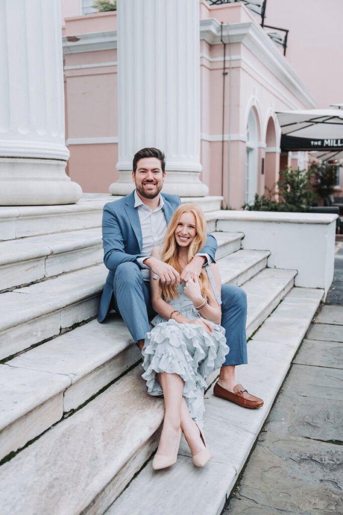 girl and guy smile towards camera as they sit on steps for their charleston engagement session